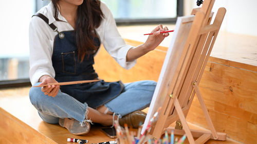 Cropped image of young artist woman drawing and sitting on the floor in front of canvas easel