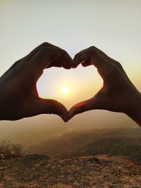 Hand holding heart shape against sky during sunset