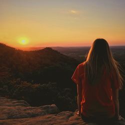 Rear view of woman sitting on mountain against sky during sunset