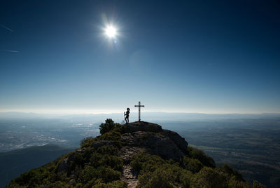 Woman contemplating a backlit landscape from the top of a hill, where there is a cross.