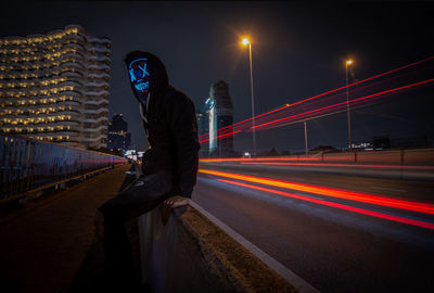 Man wearing mask sitting by light trails on bridge in city at night
