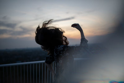 Side view of woman with arms raised standing by railing against cloudy sky during sunset