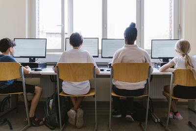 Rear view of male and female students using computers while sitting in classroom at school