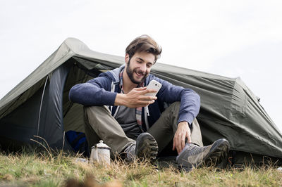 Smiling man using smart phone outside tent on hill against clear sky