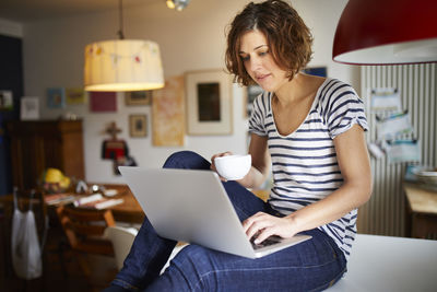 Portrait of mature woman sitting on kitchen table using laptop