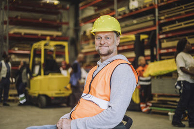 Portrait of smiling carpenter with disability wearing hardhat sitting on wheelchair in warehouse