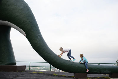 Sisters playing on statue of animal tail against clear sky