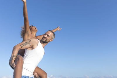 Low angle view of man piggybacking cheerful girlfriend against blue sky