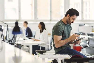 Young businessman using mobile phone while holding diary with colleagues in background at office