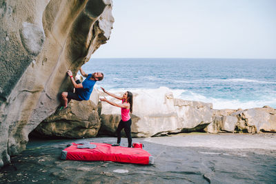 Woman looking at man climbing on rock at beach against sky