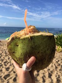 Coconut drink with straw held up to sand at beach against sky