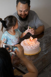 Girl photographing father and daughter with birthday cake on table