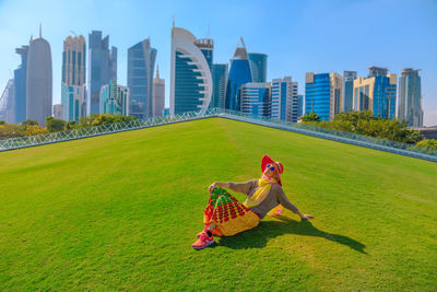 Woman sitting on field against modern buildings