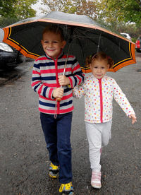 Portrait of smiling sibling holding umbrella standing on street