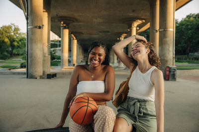 Cheerful teenage girl with basketball sitting by female friend under bridge