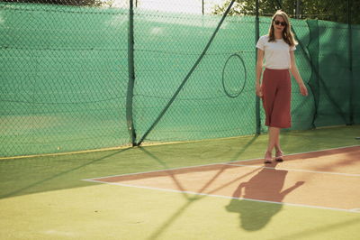 Full length portrait of woman standing on tennis court against fence