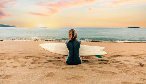 Surfer woman with wetsuit and surfboard sitting on the sand looking at the sea