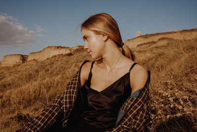 Young woman looking away while sitting by plants against sky