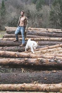 Portrait of young man standing with dog on logs of wood in forest 