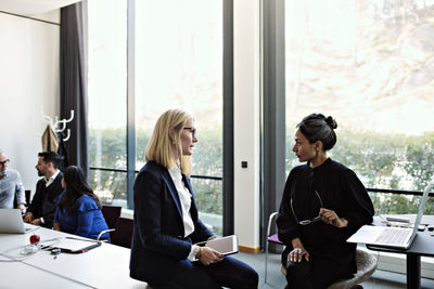 Businesswomen discussing while sitting in board room with colleagues working in background