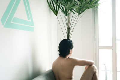 Rear view of naked man sitting on bed while looking through window