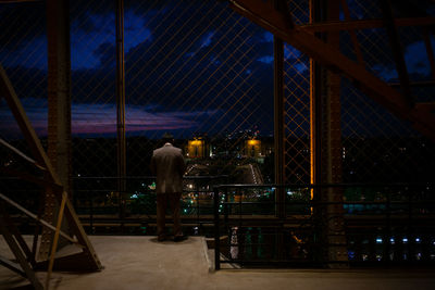 Rear view of man looking at city through fence at night