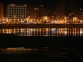 Illuminated buildings by beach against sky at night