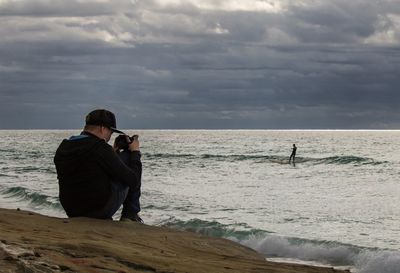 Man photographing person surfing in sea against cloudy sky