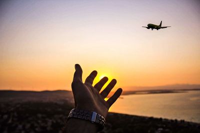 Cropped hand gesturing while airplane flying in sky during sunset