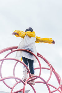 Back view of anonymous child in raincoat standing with arms apart on symmetrical construction under light sky in rainy weather