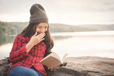 Young woman lying on rock reading book by lake against clear sky