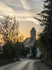 Road amidst trees and buildings against sky during sunset