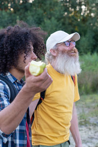 Two friends of different ages go hiking with backpacks,and an arab guy with black hair eats apple 