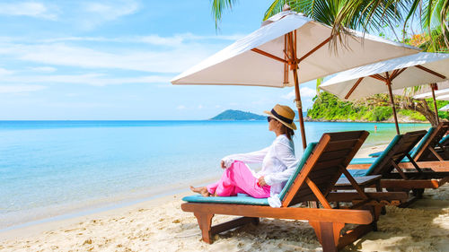 Rear view of woman sitting on lounge chairs at beach against sky