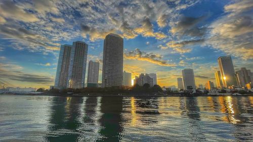 River by modern buildings against sky during sunset