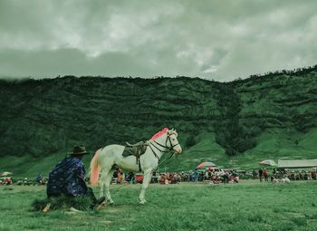 Side view of man sitting by horse on grass against mountains and cloudy sky