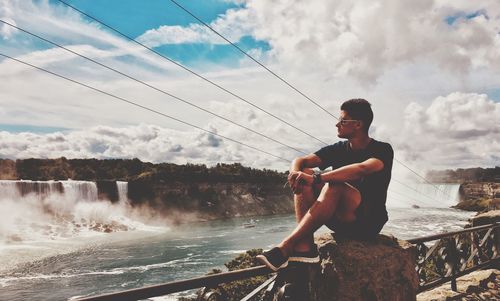 Full length of man sitting on railing by waterfall against sky