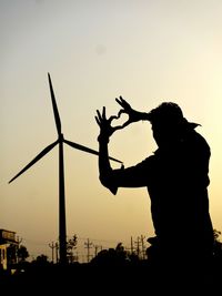 Silhouette man forming heart shape against windmill during sunset