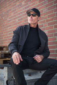 Man wearing black jacket and sunglasses while sitting against brick wall