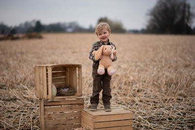A happy boy with a soft toy is standing on a box in the field