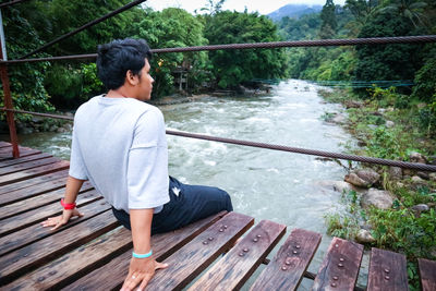 Side view of man sitting on footbridge over river in forest