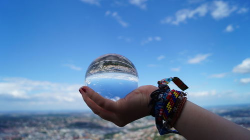 Cropped hand of woman holding crystal ball against blue sky