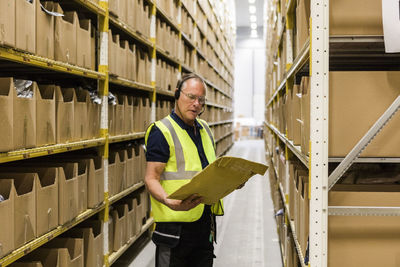 Senior male worker looking at package while talking through headset standing amidst racks at distribution warehouse