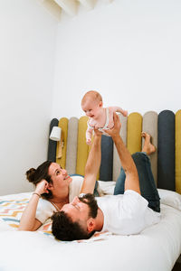 Father pretending that playful baby flying like superhero while chilling together with wife and kid on soft bed at home