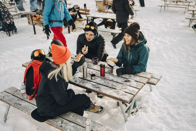 Male and female friends talking while eating hot dog at ski resort during winter