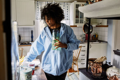 Young woman with curly hair holding drinking glasses while standing in kitchen at home