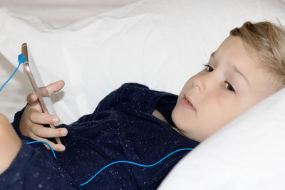 Little boy with headphones using mobile phone while lying down on bed at home.