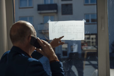 Architect talking through mobile phone analyzing blueprint on window