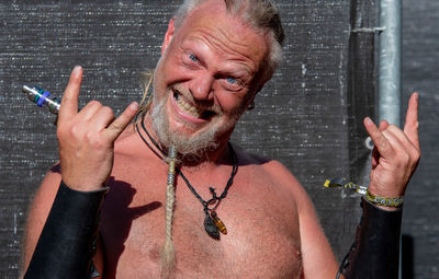 Portrait of cheerful shirtless man gesturing horn sign against wall