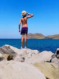Rear view of shirtless boy standing on rock at beach